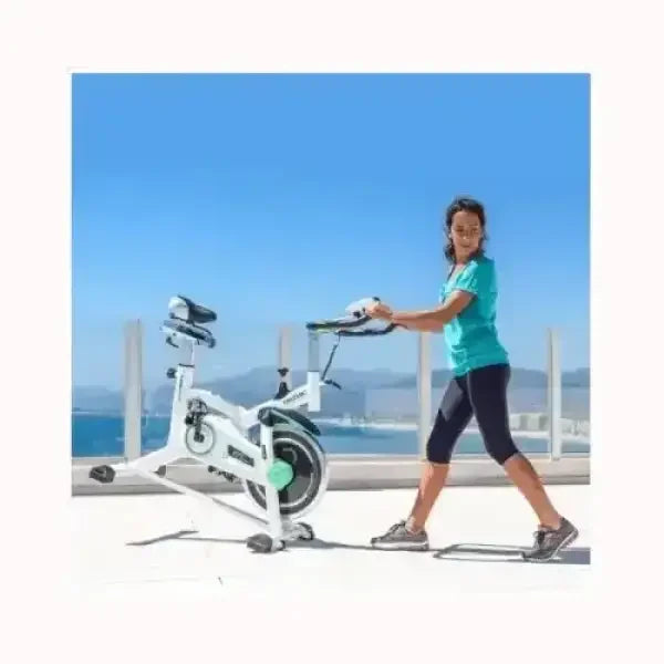 Donna che regola Cecotec Spin Bike bianca su terrazza al mare con sedile e manubrio regolabili per allenamento